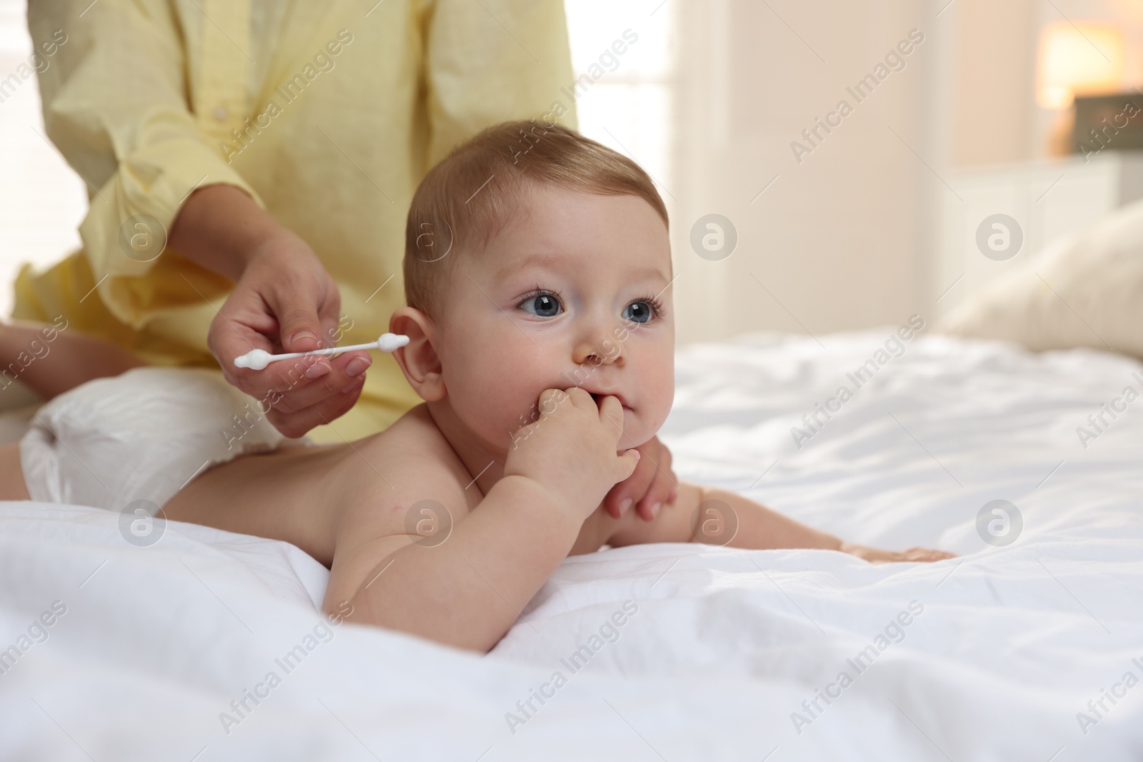 Photo of Mother cleaning ear of her cute little baby with cotton swab on bed, closeup