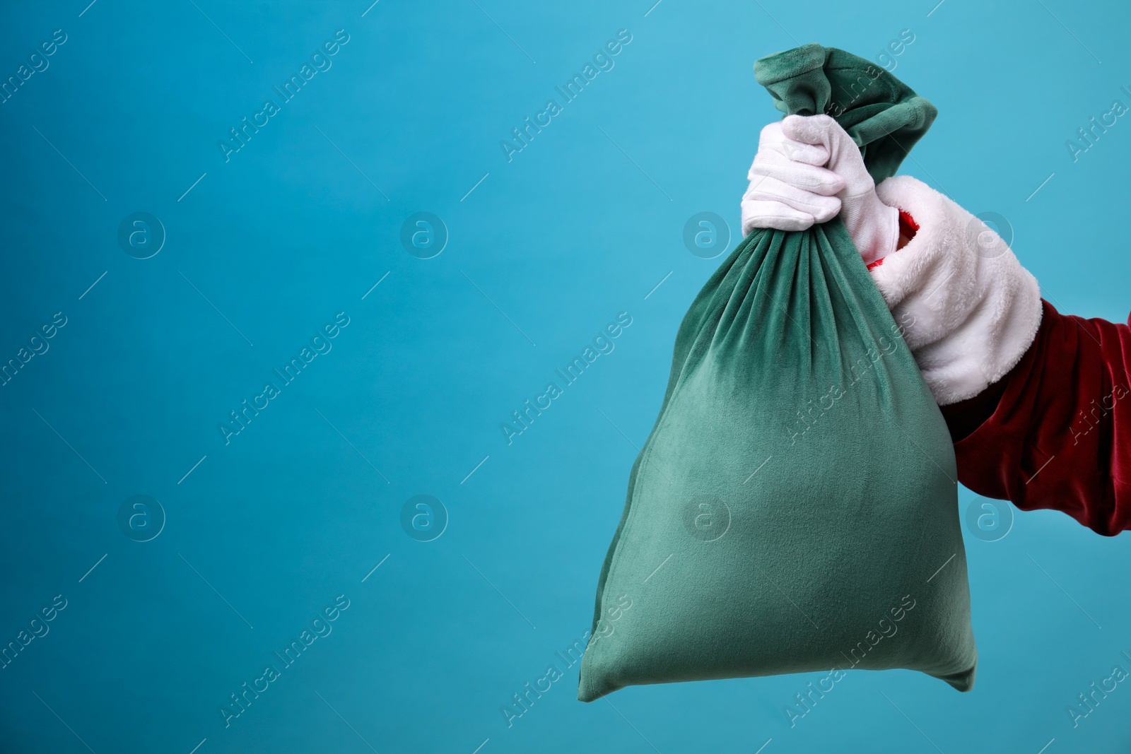 Photo of Santa Claus with green bag of Christmas gifts on light blue background, closeup. Space for text