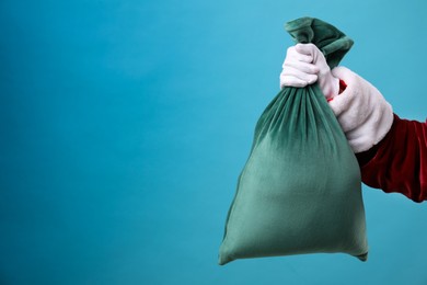 Photo of Santa Claus with green bag of Christmas gifts on light blue background, closeup. Space for text