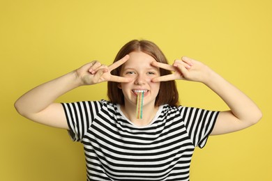 Photo of Teenage girl eating tasty rainbow sour belt while showing v-sign on yellow background