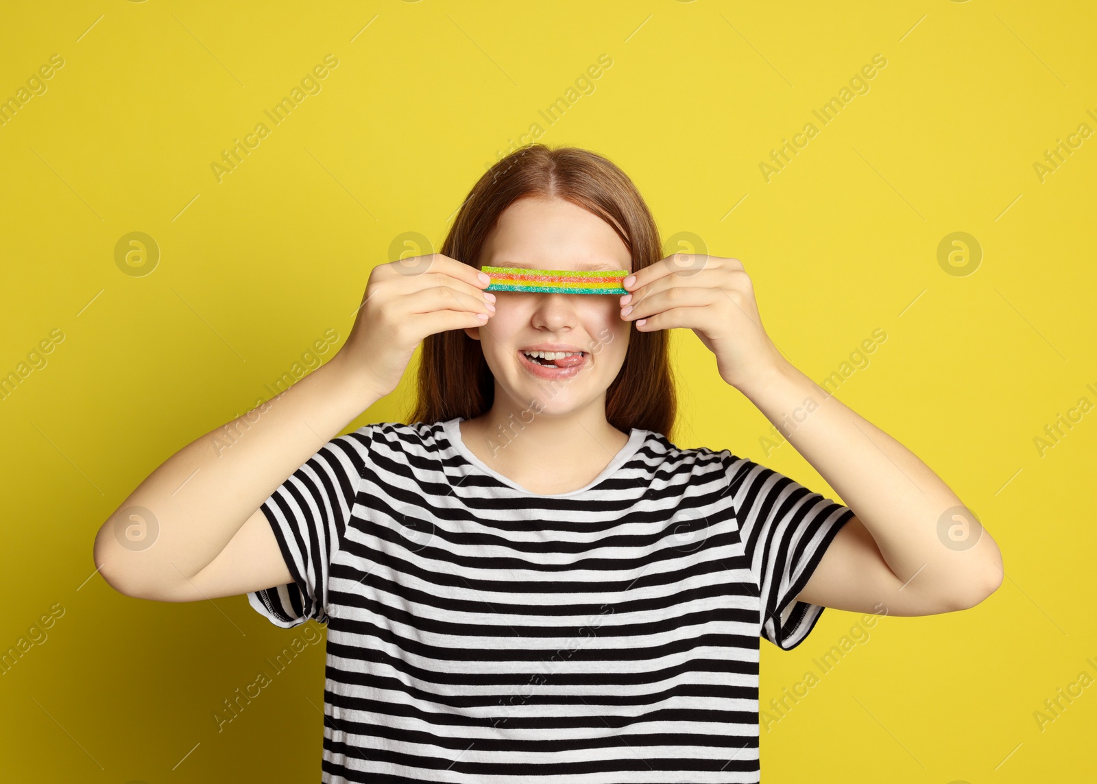 Photo of Happy teenage girl with tasty rainbow sour belt closing her eyes on yellow background