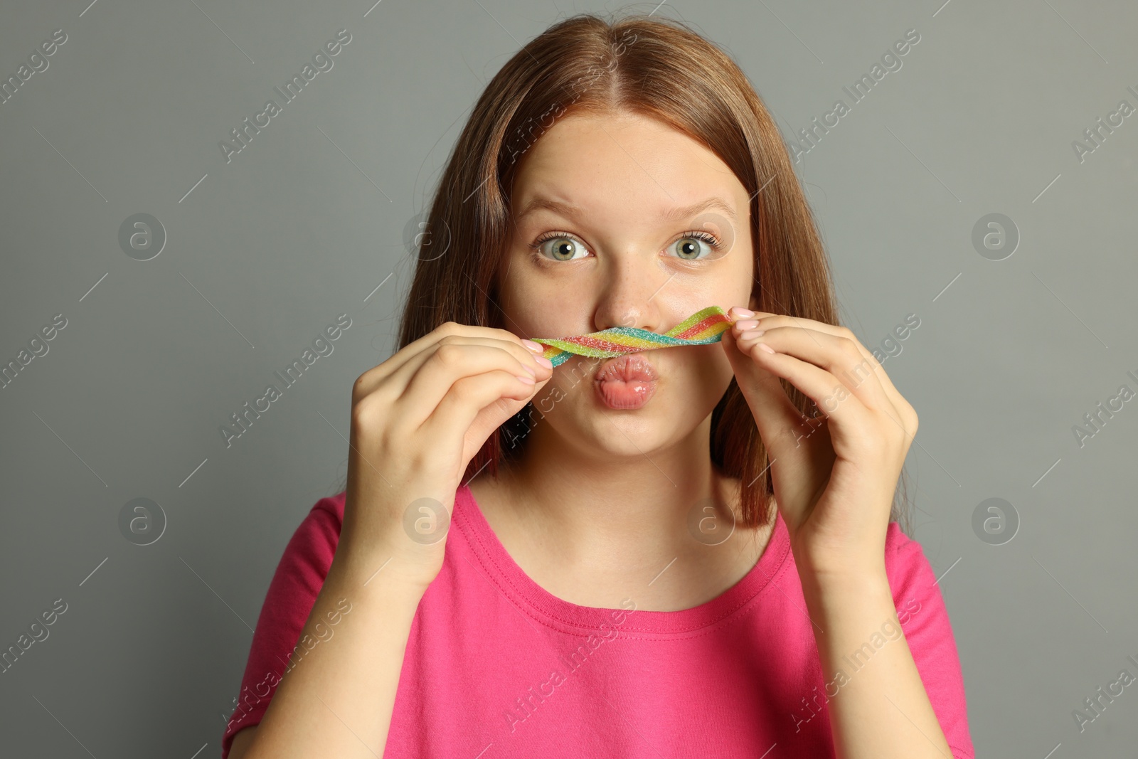 Photo of Funny teenage girl with tasty rainbow sour belt on grey background