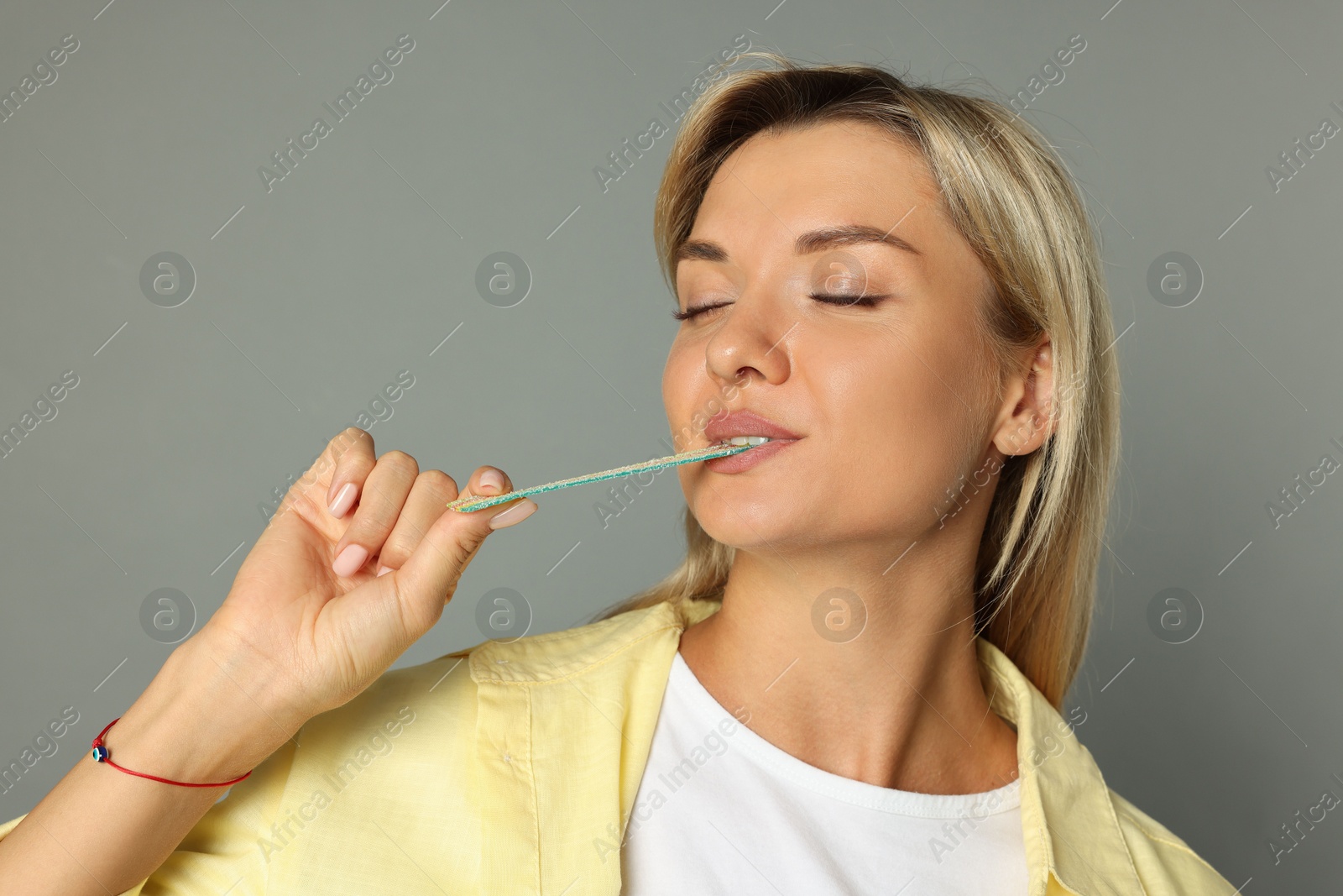 Photo of Young woman eating tasty rainbow sour belt on grey background