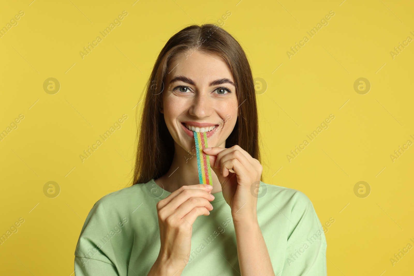 Photo of Young woman eating tasty rainbow sour belt on yellow background