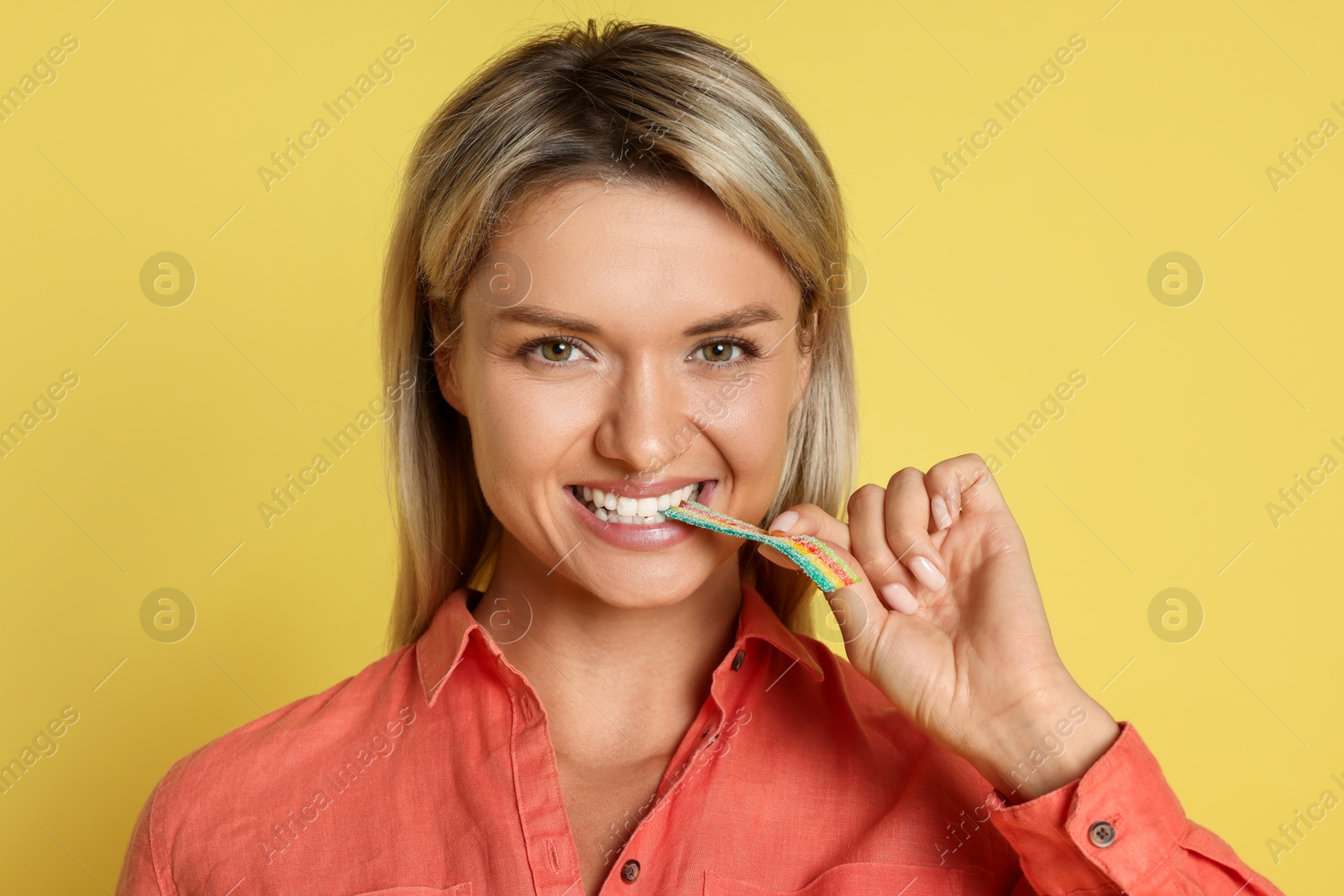 Photo of Young woman eating tasty rainbow sour belt on yellow background