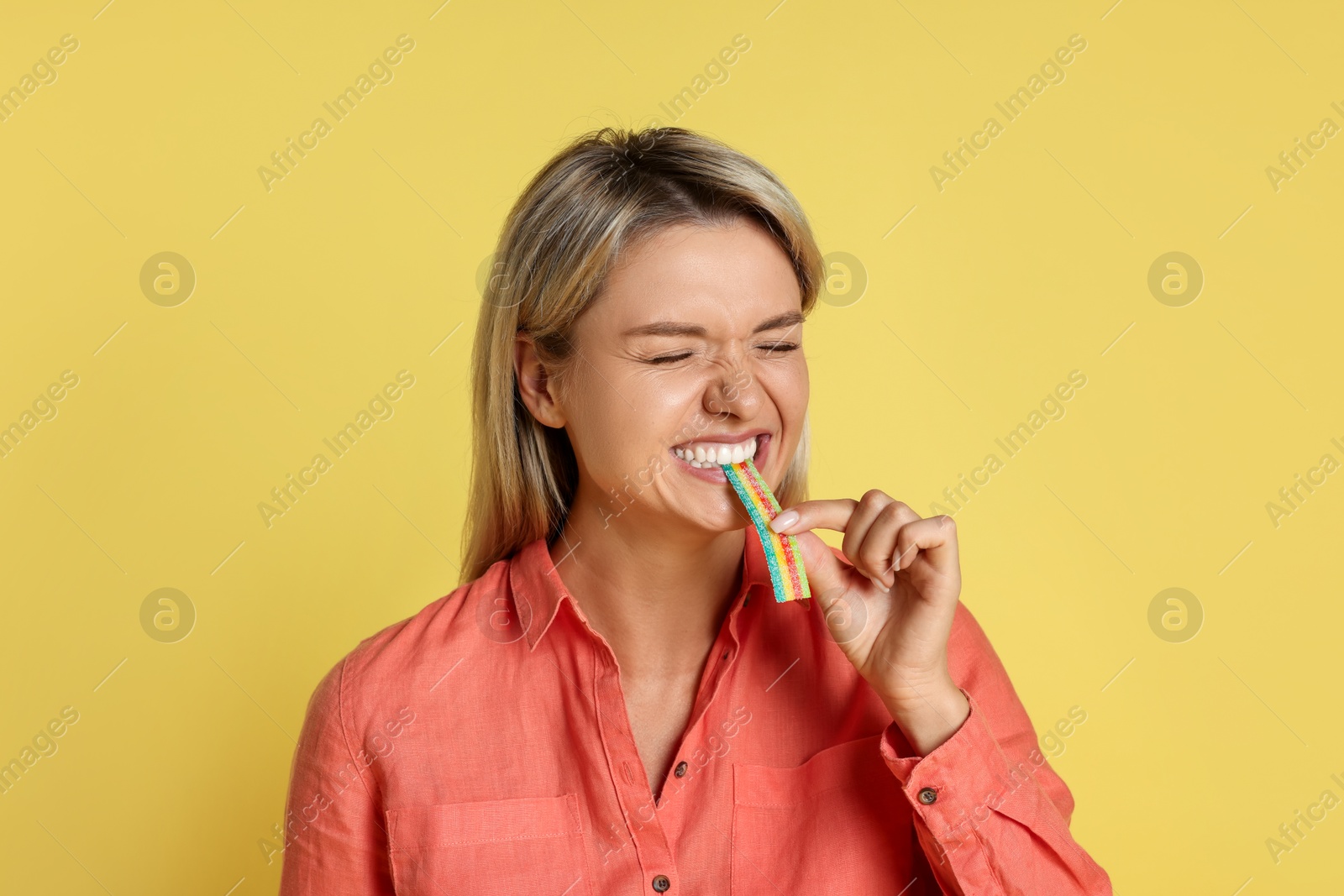 Photo of Young woman eating tasty rainbow sour belt on yellow background