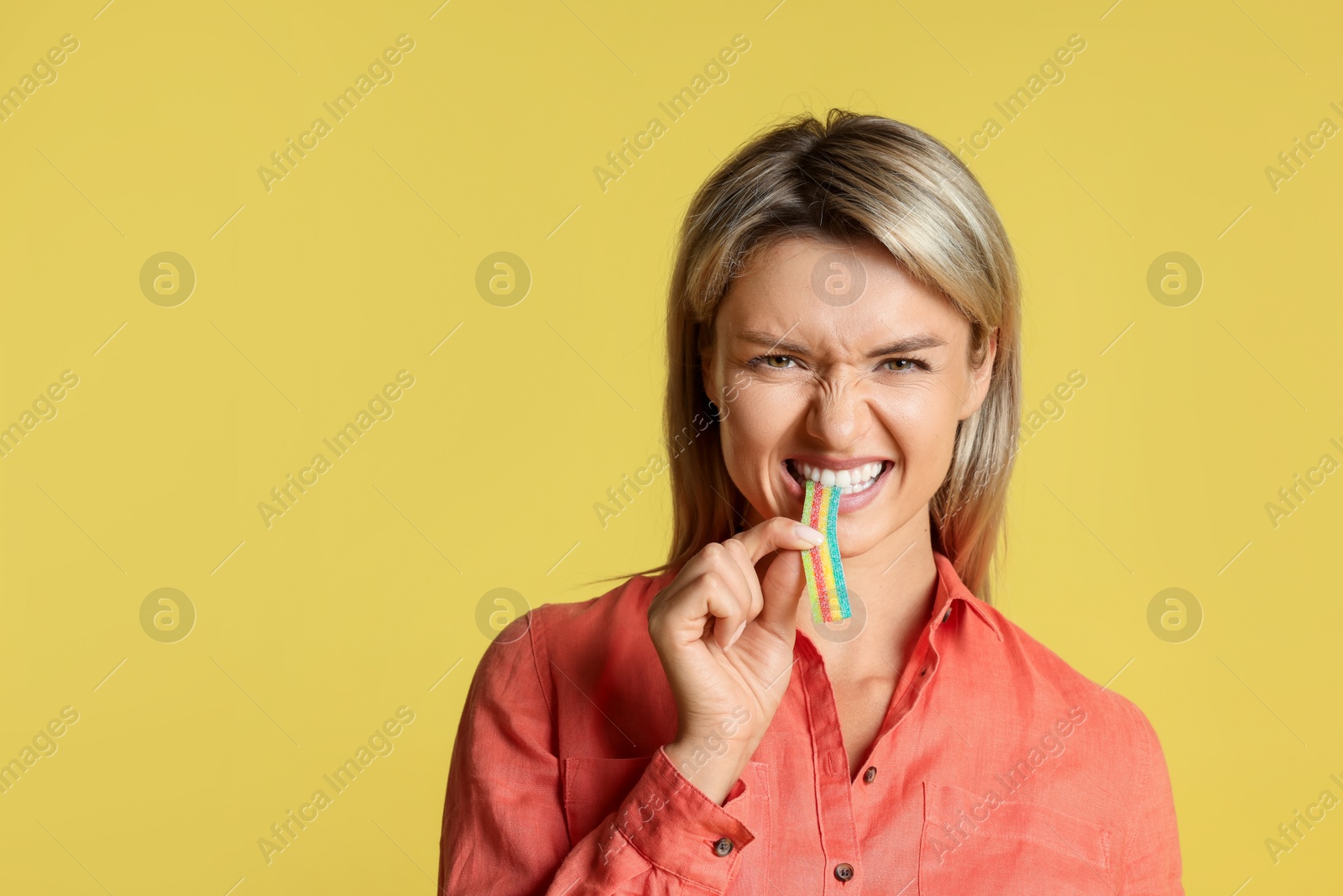 Photo of Young woman eating tasty rainbow sour belt on yellow background