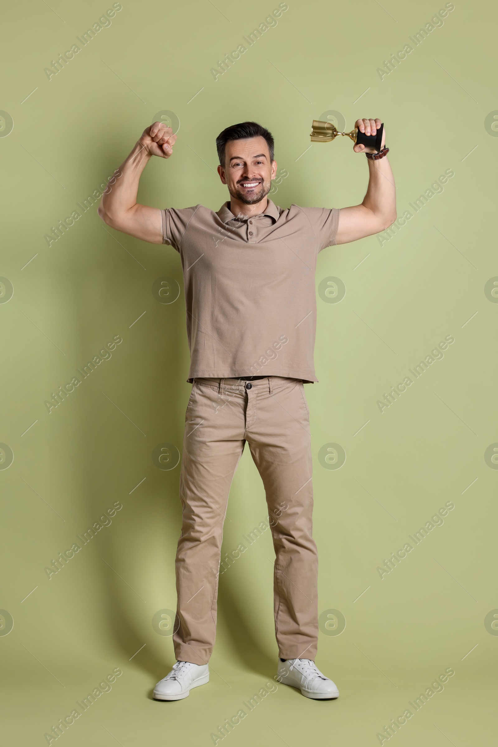 Photo of Happy winner with golden trophy cup showing his biceps on pale olive background