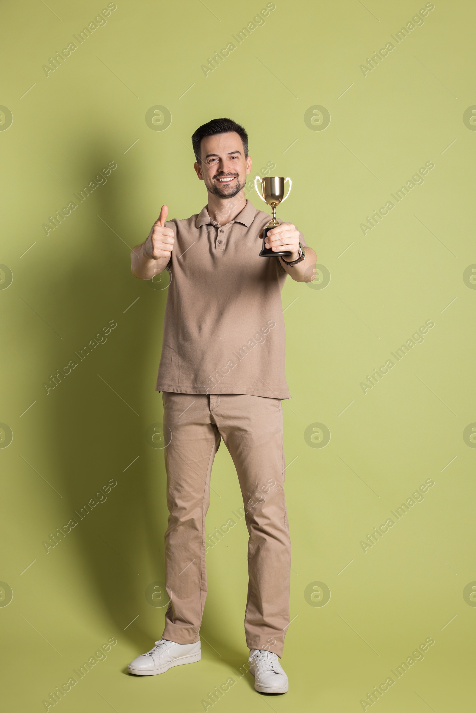 Photo of Happy winner with golden trophy cup showing thumbs up on pale olive background