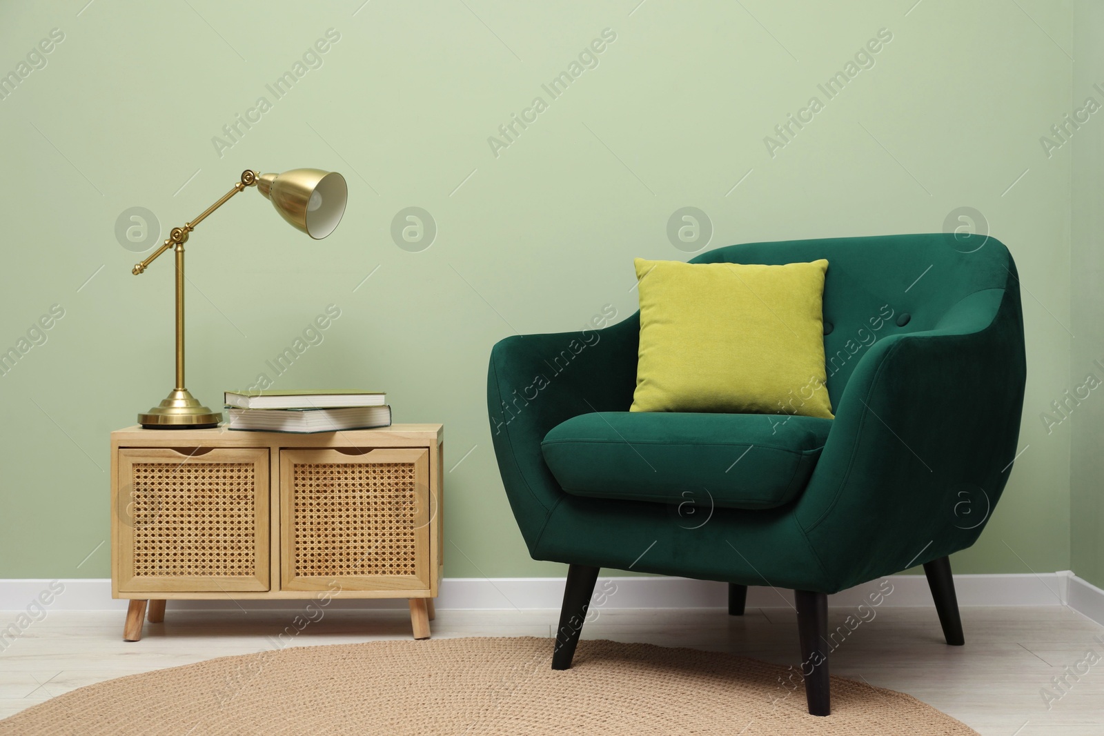 Photo of Stylish green armchair with pillow, golden lamp and books on side table near olive wall