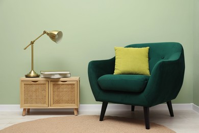 Photo of Stylish green armchair with pillow, golden lamp and books on side table near olive wall