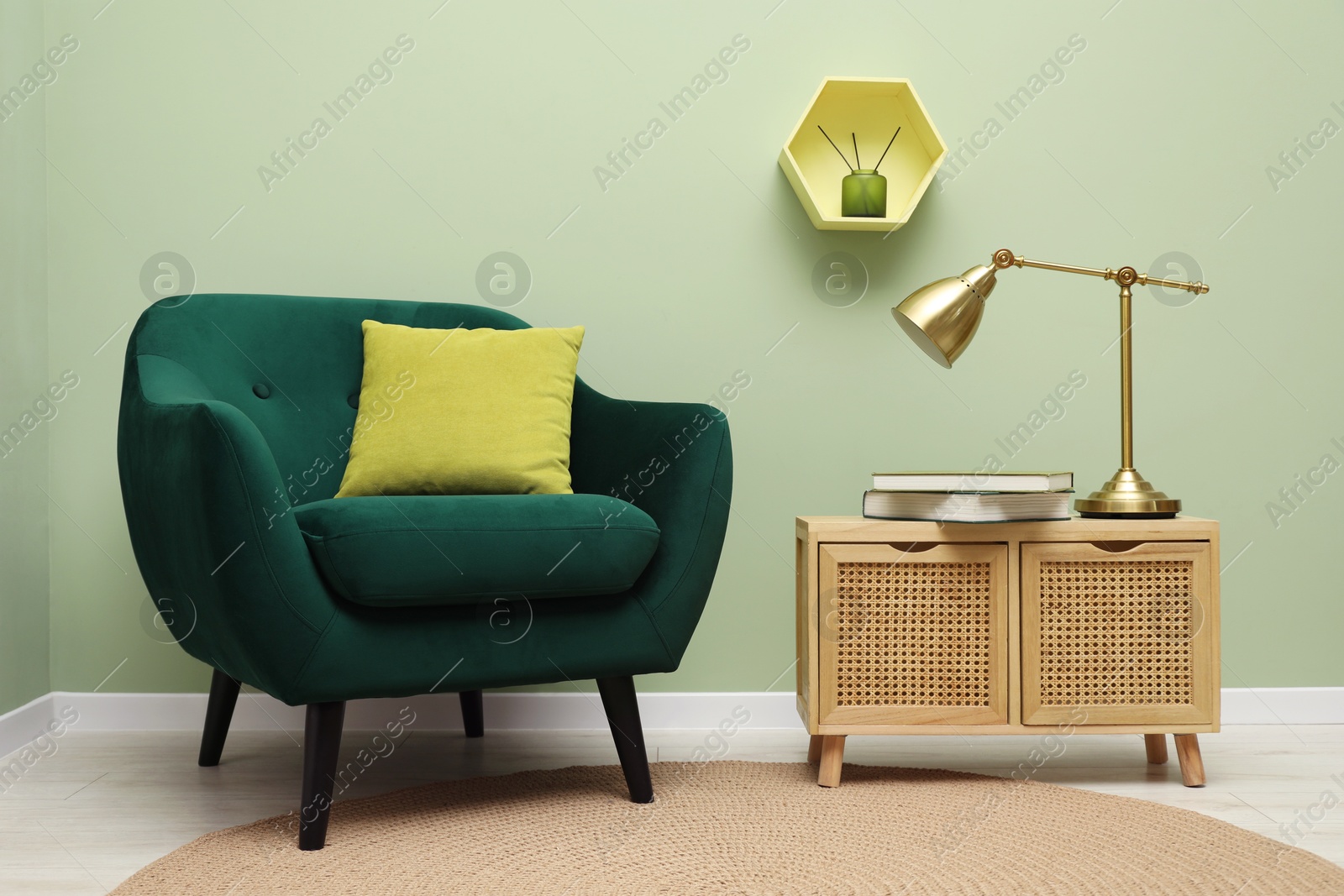 Photo of Stylish green armchair with pillow, golden lamp and books on side table near olive wall