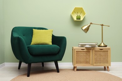 Photo of Stylish green armchair with pillow, golden lamp and books on side table near olive wall