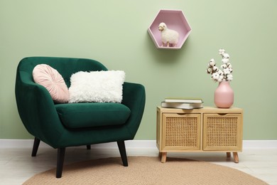 Photo of Stylish green armchair with soft pillows, cotton flowers in vase and books on side table near olive wall