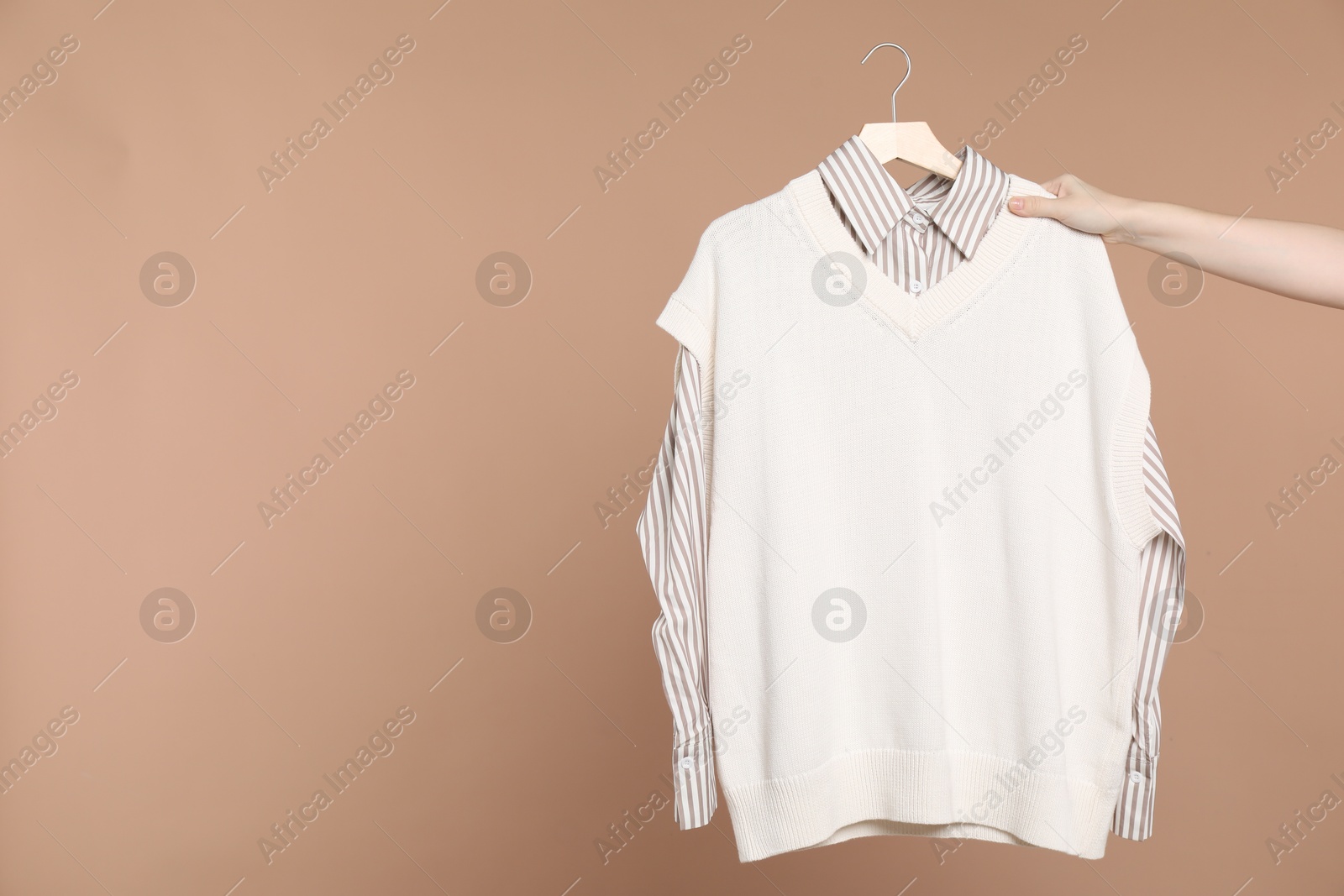 Photo of Woman holding hanger with shirt and sweater vest on beige background, closeup. Space for text