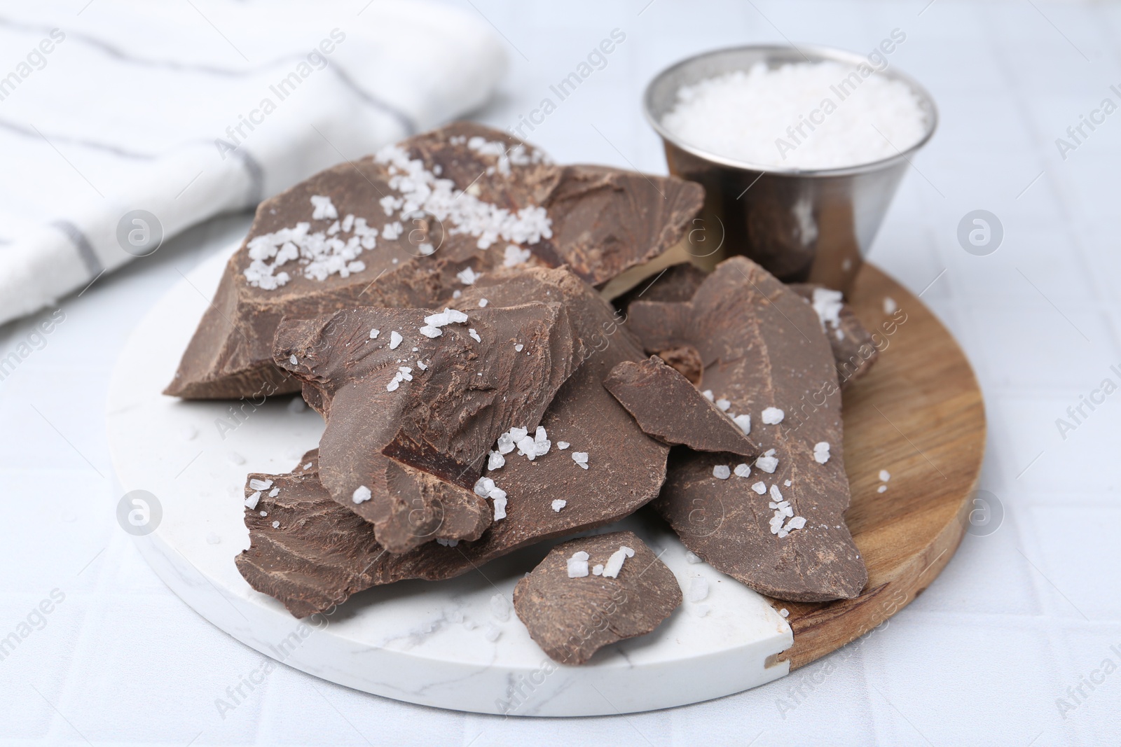 Photo of Pieces of tasty chocolate with salt on white tiled table, closeup