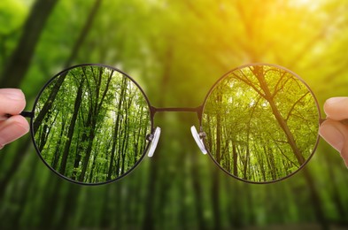 Image of Woman holding glasses against in forest, closeup. View through lenses more clear. Vision correction