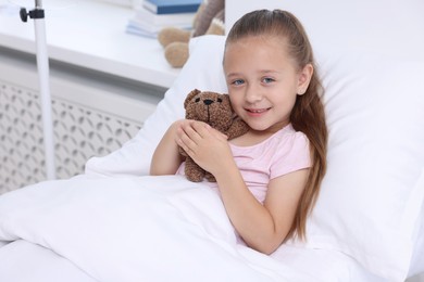 Photo of Cute little girl with teddy bear on bed in hospital