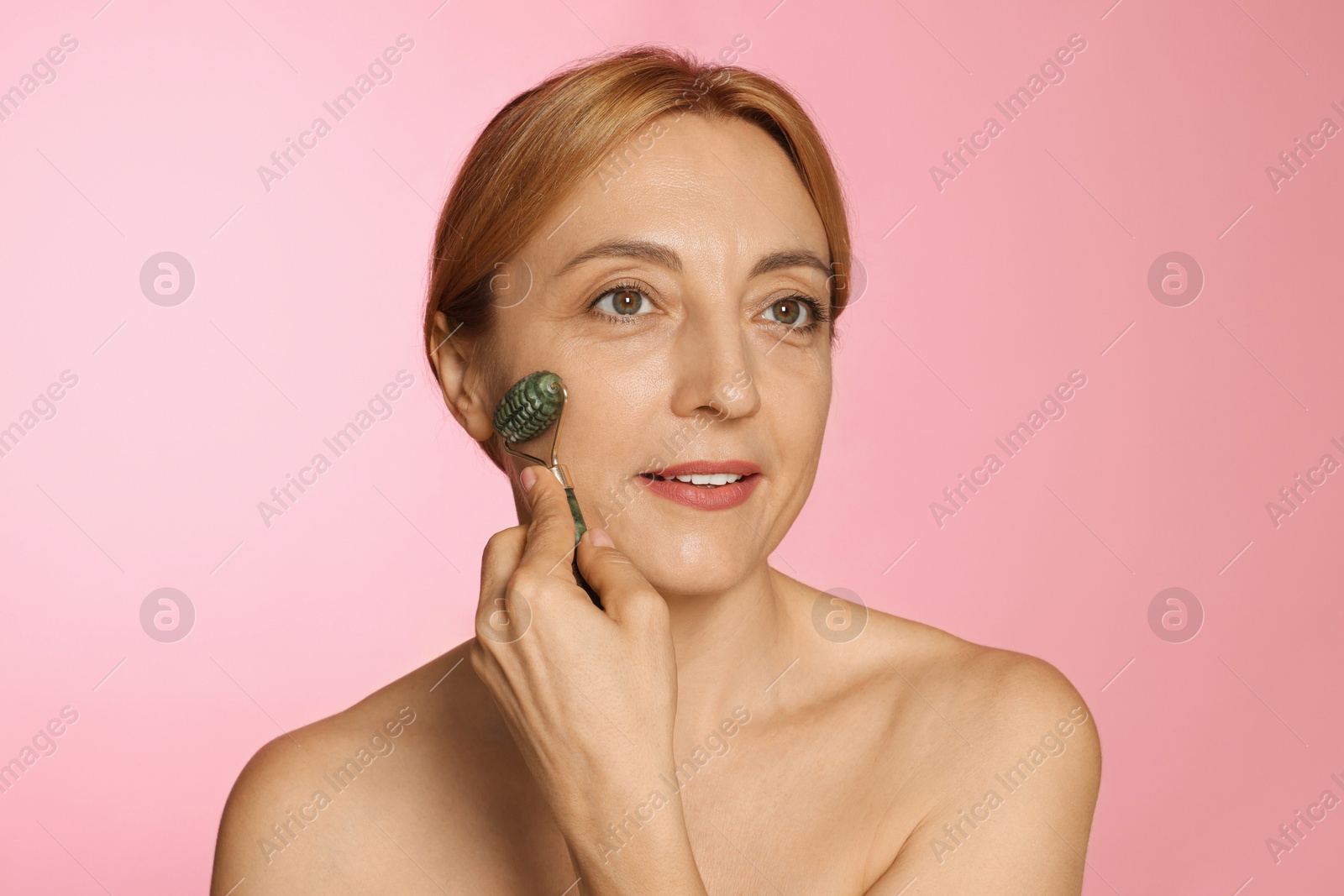 Photo of Beautiful woman doing facial self massage with roller on pink background