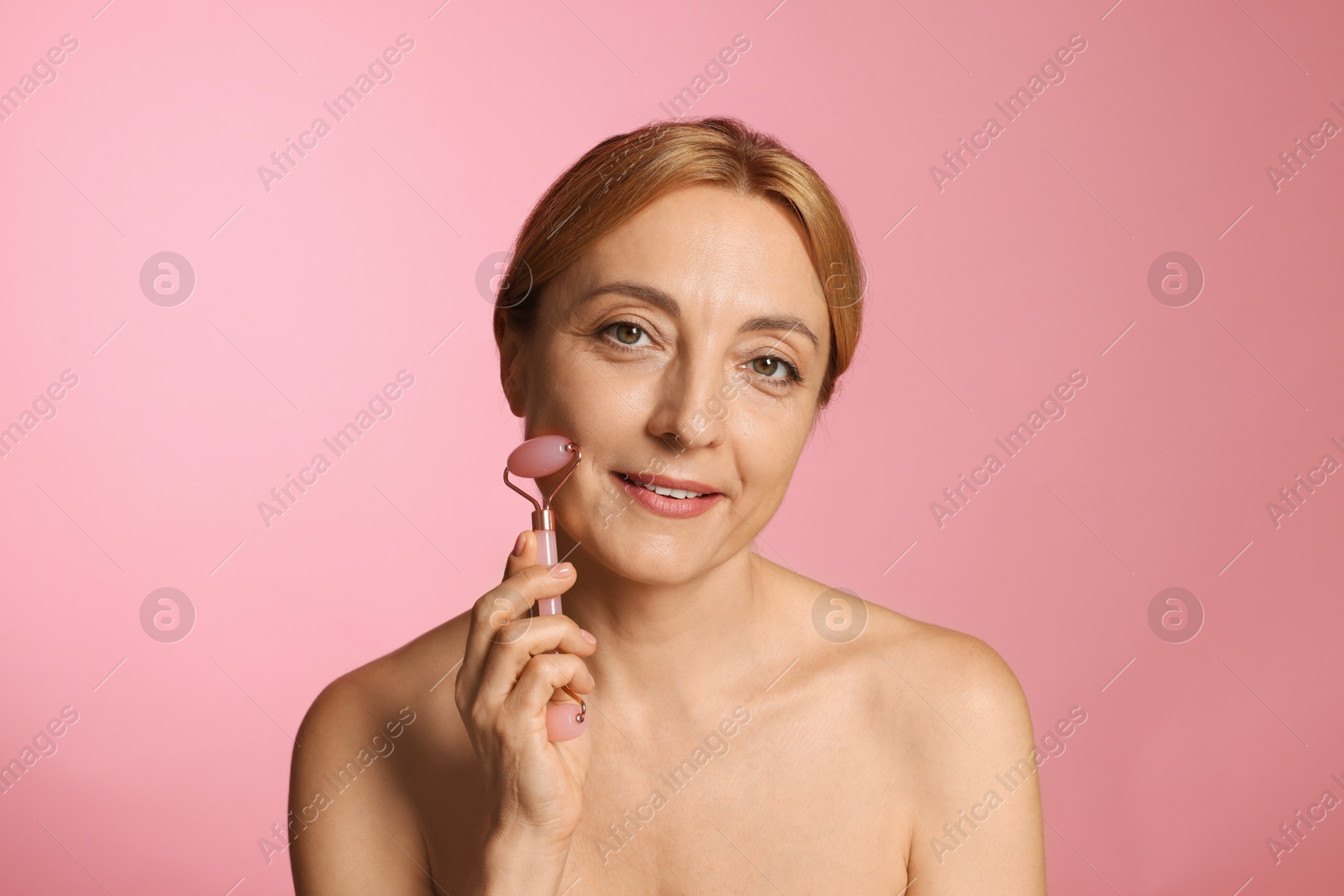 Photo of Smiling woman doing facial self massage with roller on pink background