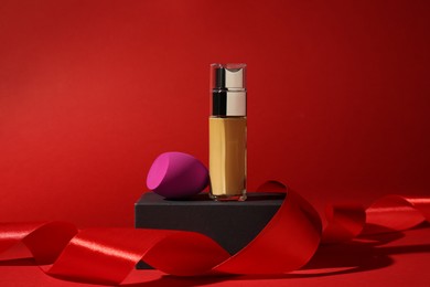 Photo of Christmas present. Liquid foundation, sponge and gift box on red background