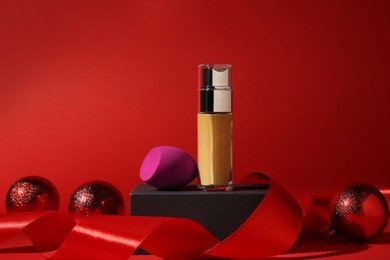 Photo of Christmas present. Liquid foundation, sponge, gift box and festive decor on red background