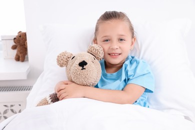 Photo of Cute little girl with teddy bear on bed in hospital