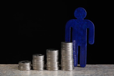 Stacked coins and paper human figure on wooden table against black background, closeup. Salary concept