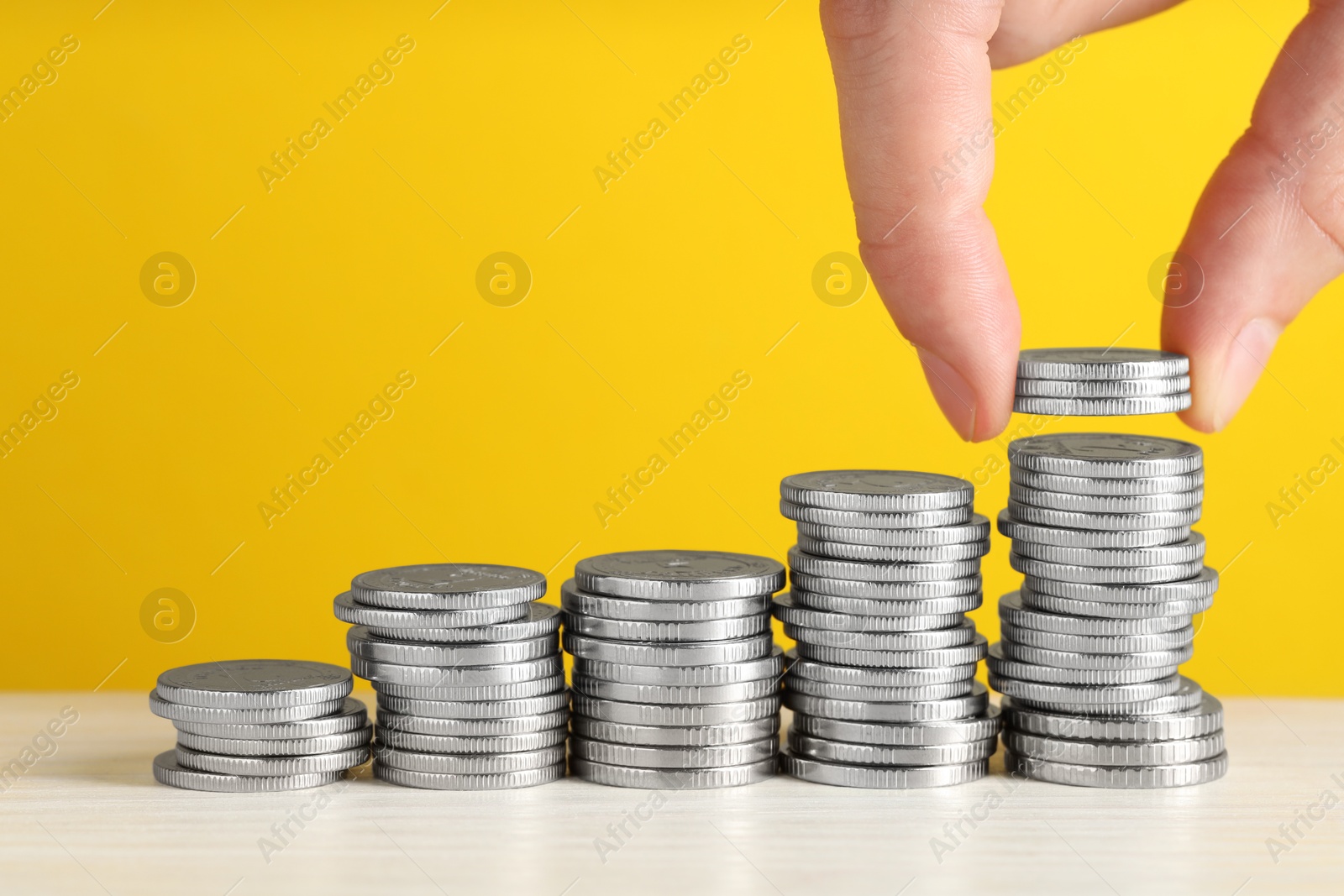 Photo of Woman putting coins at white wooden table against yellow background, closeup. Salary concept