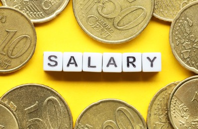 Photo of Word Salary made of cubes and coins on yellow background, flat lay