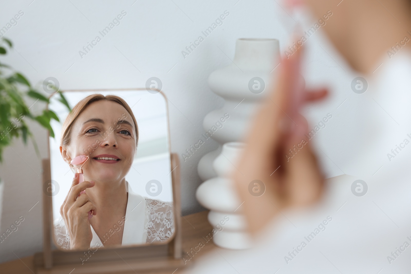 Photo of Smiling woman doing facial self massage with roller near mirror at home