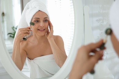 Photo of Smiling woman doing facial self massage with roller near mirror in bathroom