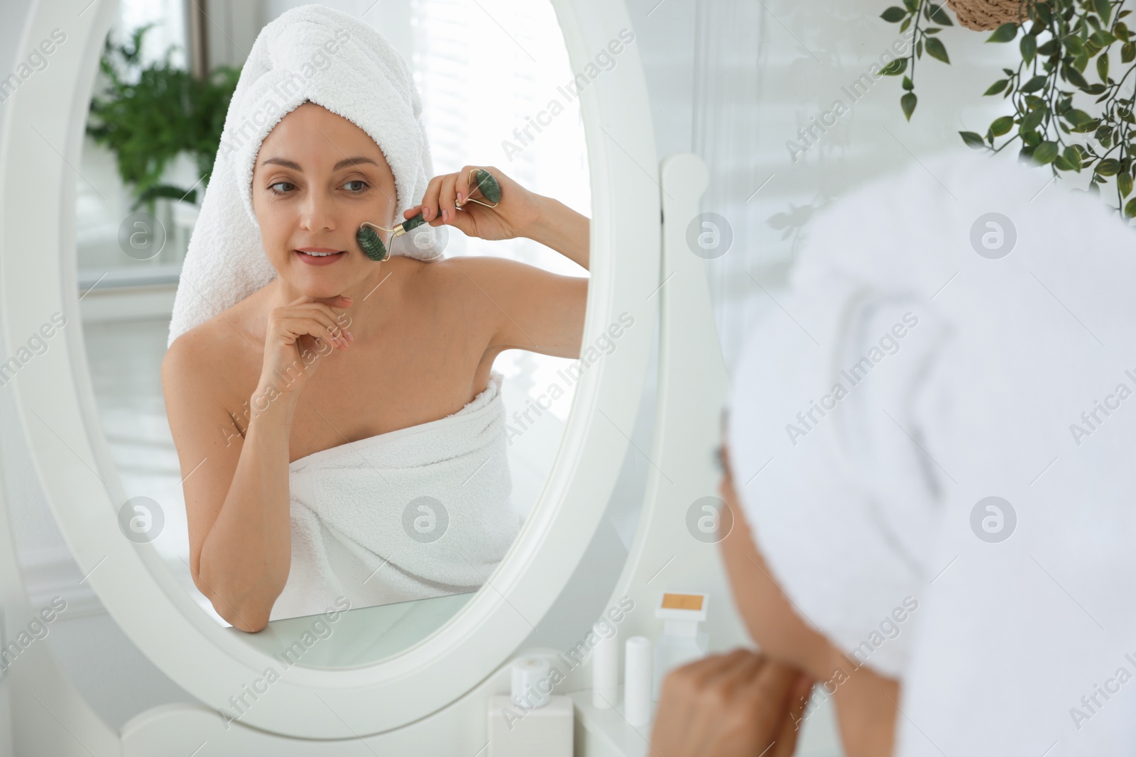 Photo of Smiling woman doing facial self massage with roller near mirror in bathroom