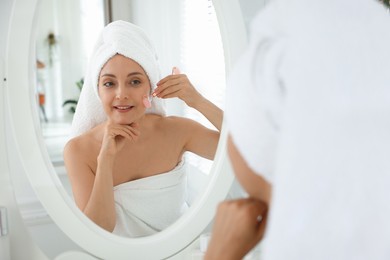 Photo of Smiling woman doing facial self massage with roller near mirror in bathroom