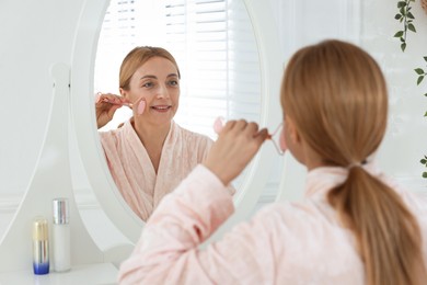 Smiling woman doing facial self massage with roller near mirror in bathroom