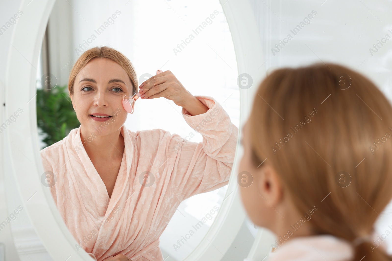 Photo of Smiling woman doing facial self massage with roller near mirror in bathroom