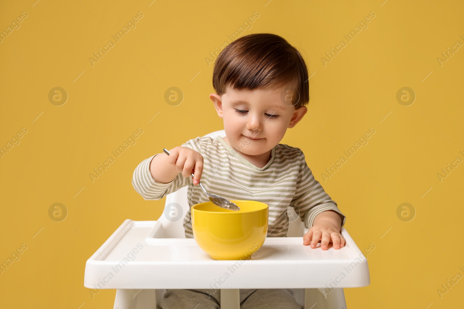 Photo of Cute little kid eating healthy baby food from bowl in high chair on yellow background