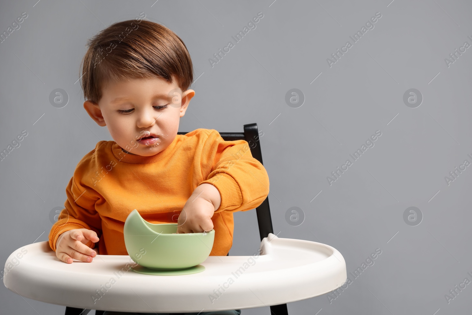 Photo of Cute little kid eating healthy baby food from bowl in high chair on light grey background