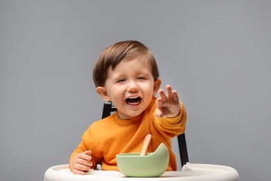 Cute little kid eating healthy baby food from bowl in high chair on light grey background