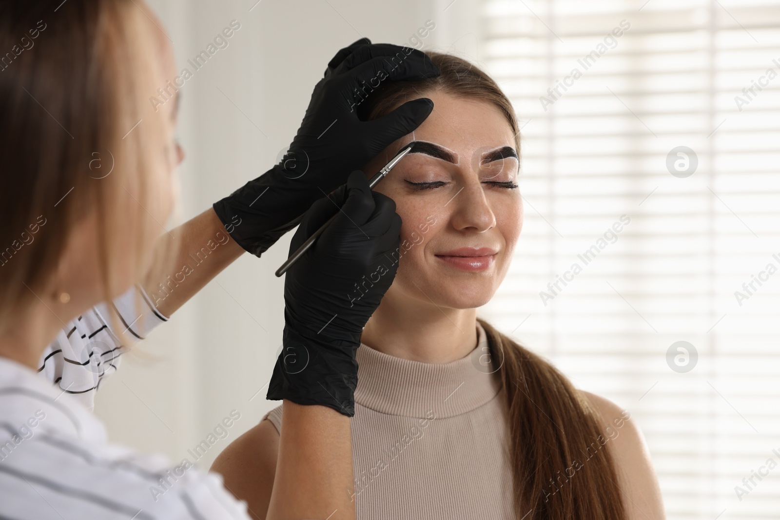 Photo of Beautician dyeing client’s eyebrows with henna in salon, closeup