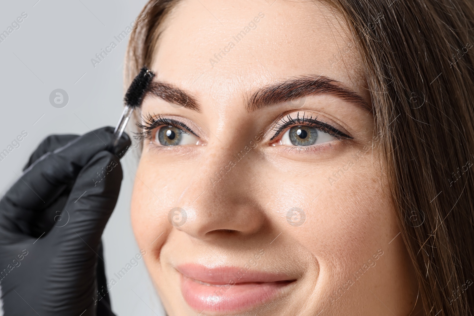 Photo of Beautician brushing client's eyebrows after henna dyeing procedure on light background, closeup