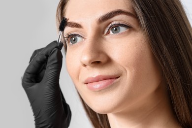 Photo of Beautician brushing client's eyebrows after henna dyeing procedure on light background, closeup