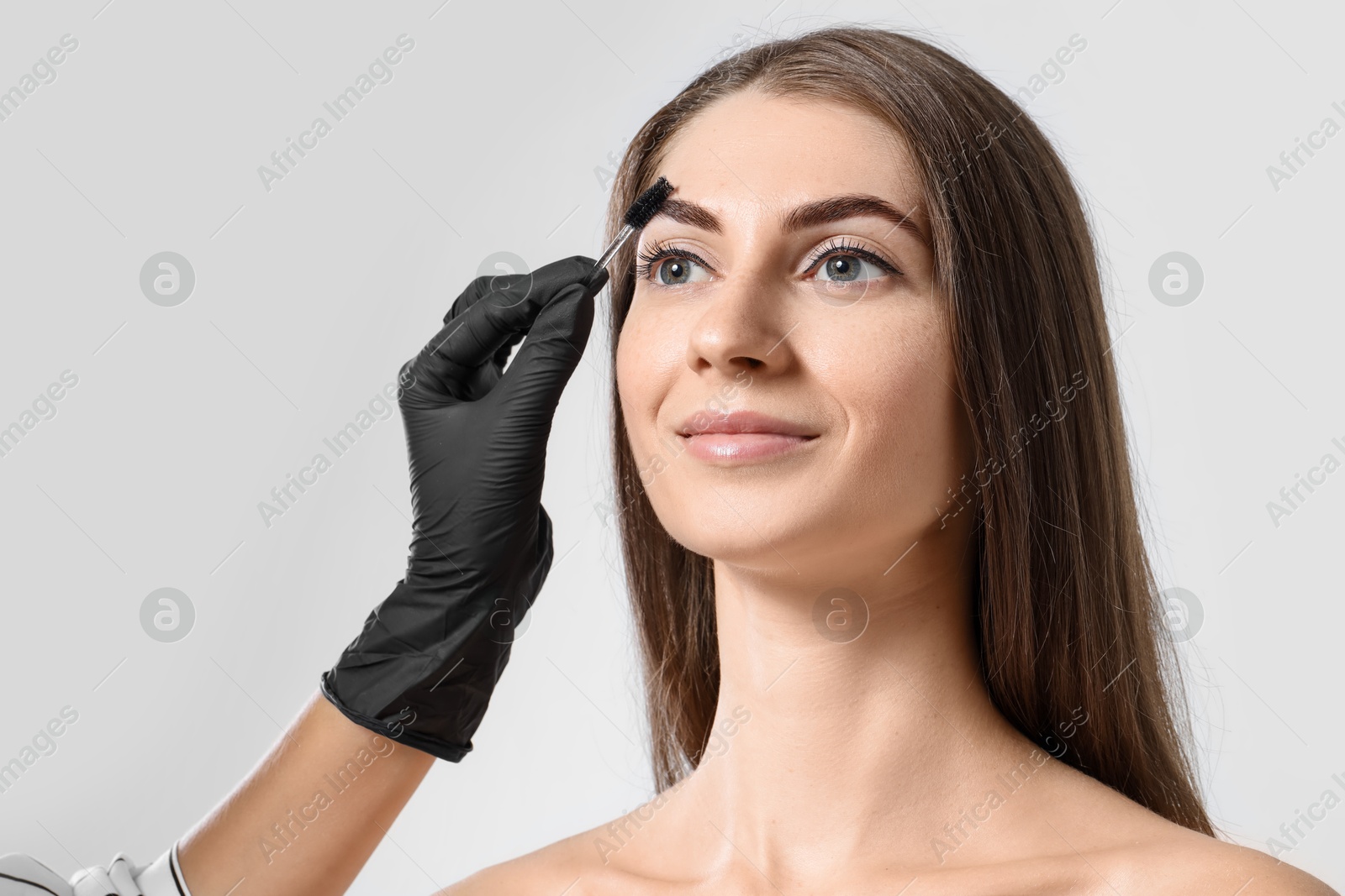 Photo of Beautician brushing client's eyebrows after henna dyeing procedure on light background, closeup