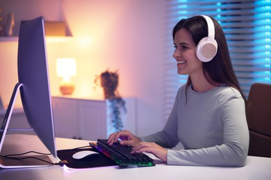 Photo of Smiling woman in headphones playing video game with keyboard at table indoors