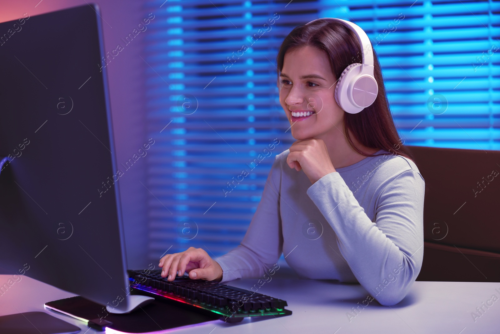 Photo of Smiling woman in headphones playing video game with keyboard at table indoors