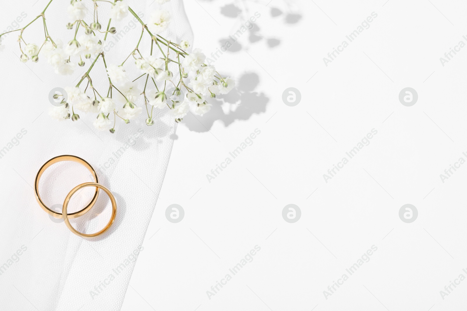 Photo of Golden wedding rings, veil and flowers on white background, top view. Space for text