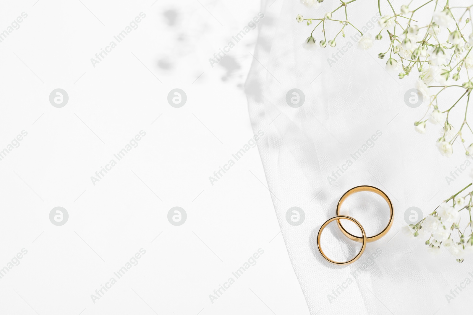 Photo of Golden wedding rings, veil and flowers on white background, top view. Space for text