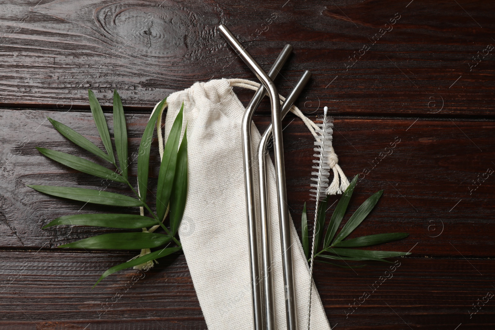 Photo of Metal drinking straws, cleaning brush, palm leaves and bag on wooden table, flat lay