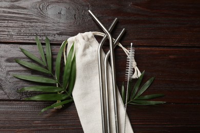 Photo of Metal drinking straws, cleaning brush, palm leaves and bag on wooden table, flat lay