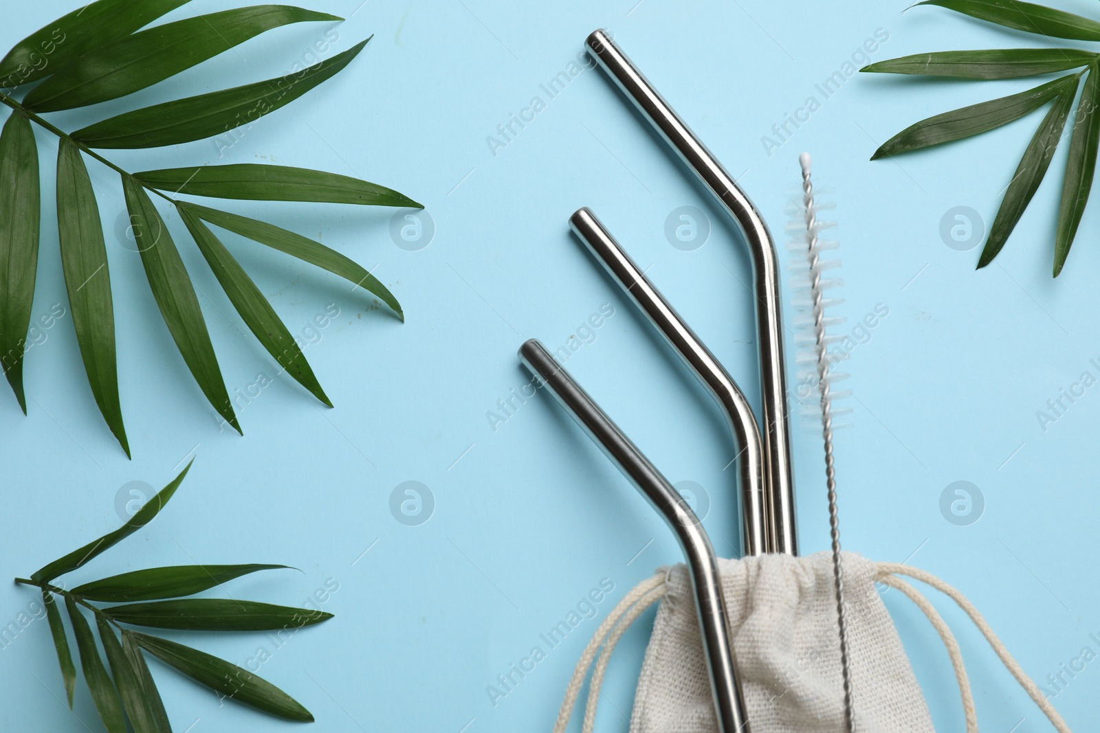 Photo of Metal drinking straws, cleaning brush, bag and palm leaves on light blue background, flat lay